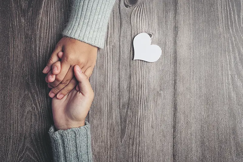 Two hands holding each other on wooden background with paper heart cutout